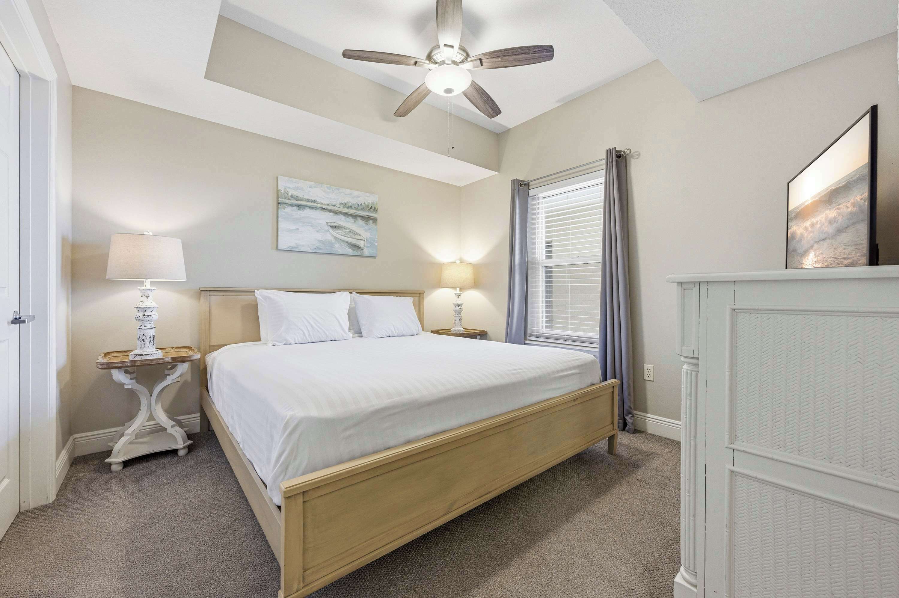 A bedroom with a bed, two bedside tables, a ceiling fan, a window with curtains, and a wall-mounted picture.