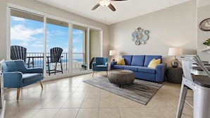 A spacious living room with a large sliding glass door leading to a balcony, a ceiling fan, a blue sofa, two armchairs, and a coffee table. - 20th Floor Condo With Stunning View (Miramar Beach)