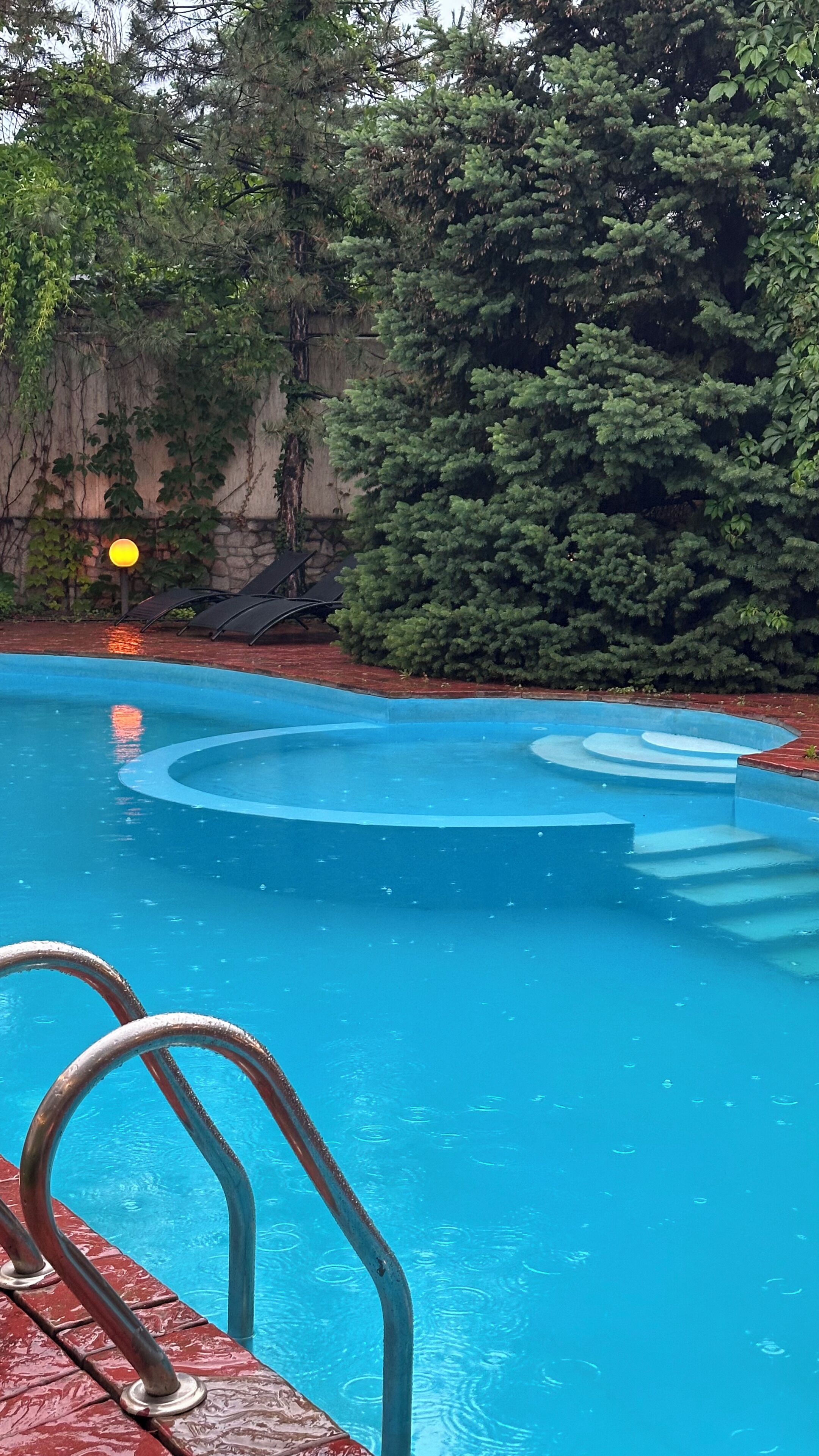 Une piscine dotée d’une plage rouge, entourée d’arbres et d’un mur de pierre.
