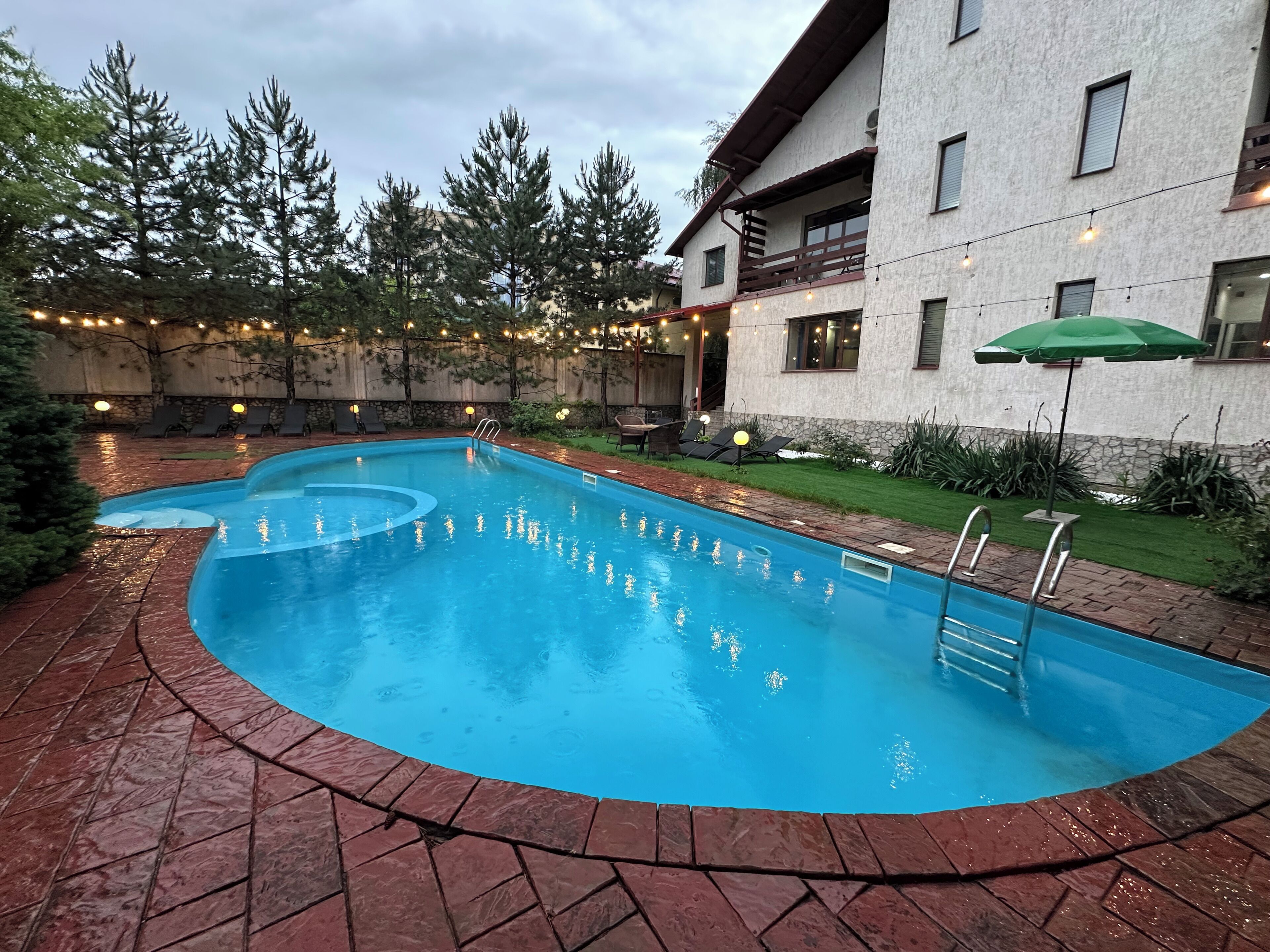 A swimming pool with a curved edge, surrounded by a brick patio. There are lounge chairs and a green umbrella.