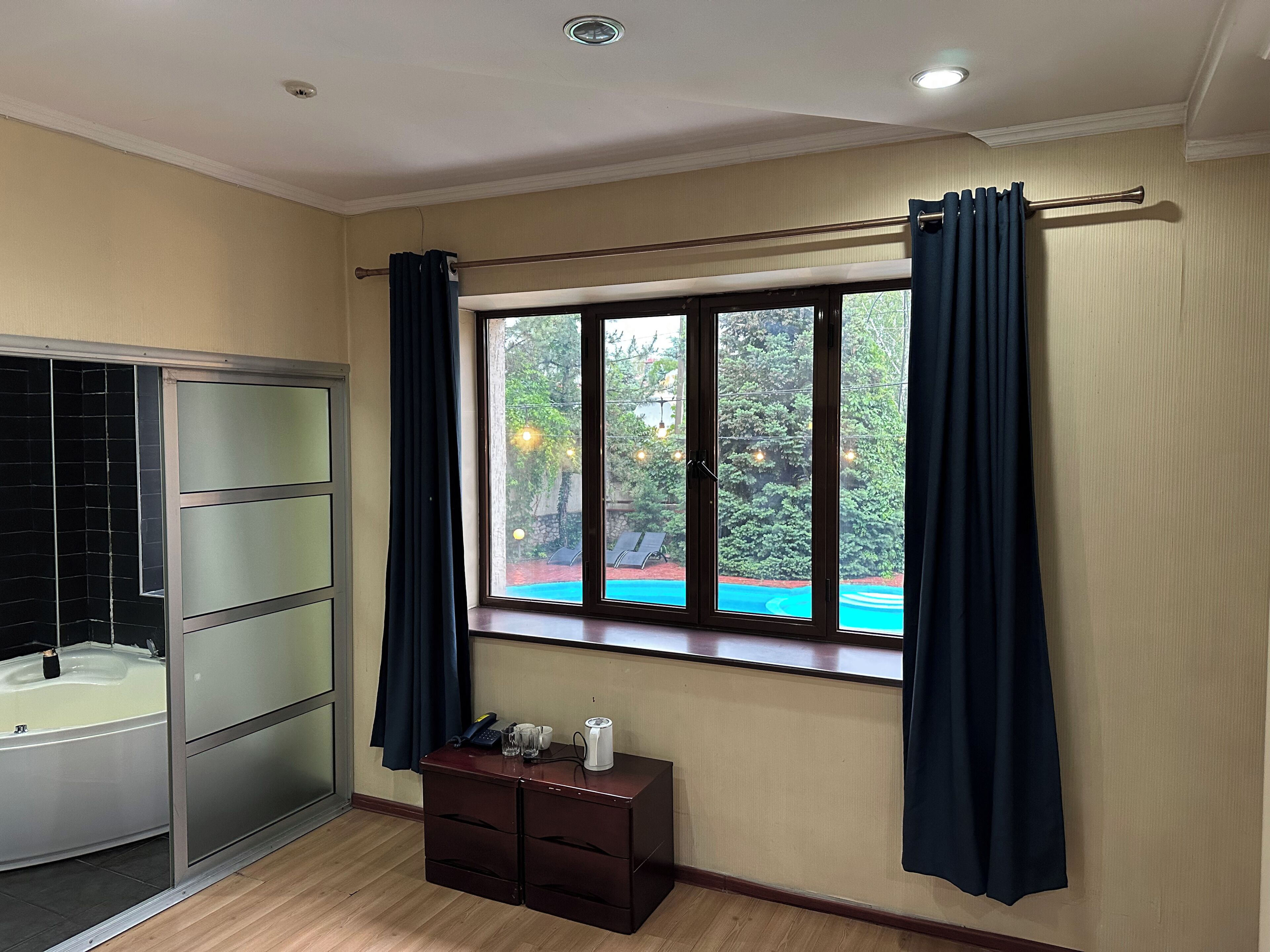 Un baño con bañera, una ventana con cortinas y un pequeño armario de madera con artículos de aseo.