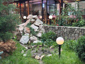 A stone wall with greenery, a wooden table with a red checkered tablecloth, and a glass building with a reflection of a person. - Gatsby Ortosay (Bishkek)