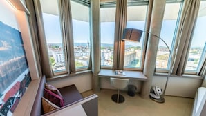 A corner office with a desk, chair, lamp, and a view of a cityscape through large windows. - aquaTurm Hotel GmbH (Radolfzell am Bodensee)