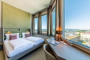 A hotel room with a bed, a desk, a chair, and a large window with a city view. - aquaTurm Hotel GmbH (Radolfzell am Bodensee)