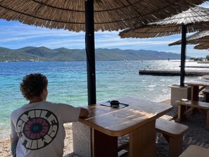 Escena costera con una mesa de madera y bancos, una sombrilla de paja y vistas al mar y colinas distantes.