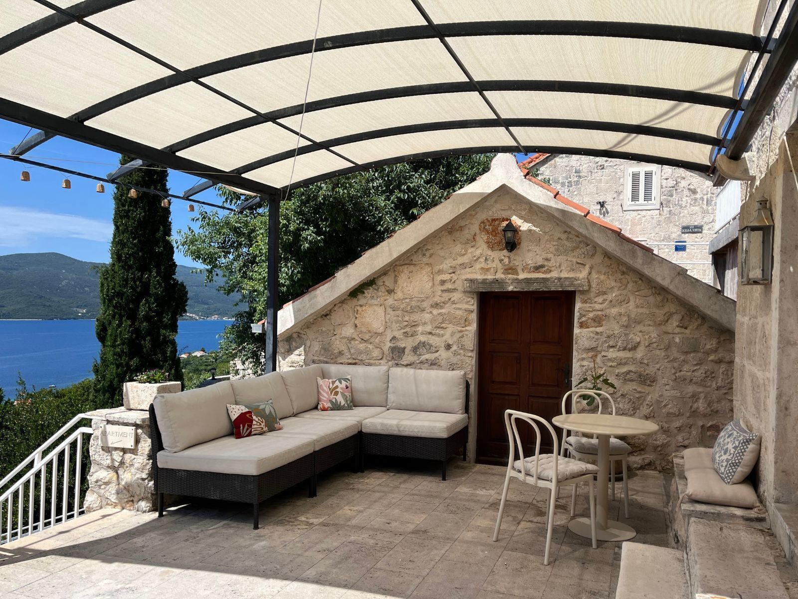 A covered outdoor seating area with a stone wall, a wooden door, and a view of the sea.