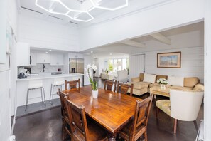 Een ruime keuken met witte kasten, een eethoek met een houten tafel, en een woonkamer met een beige bank.