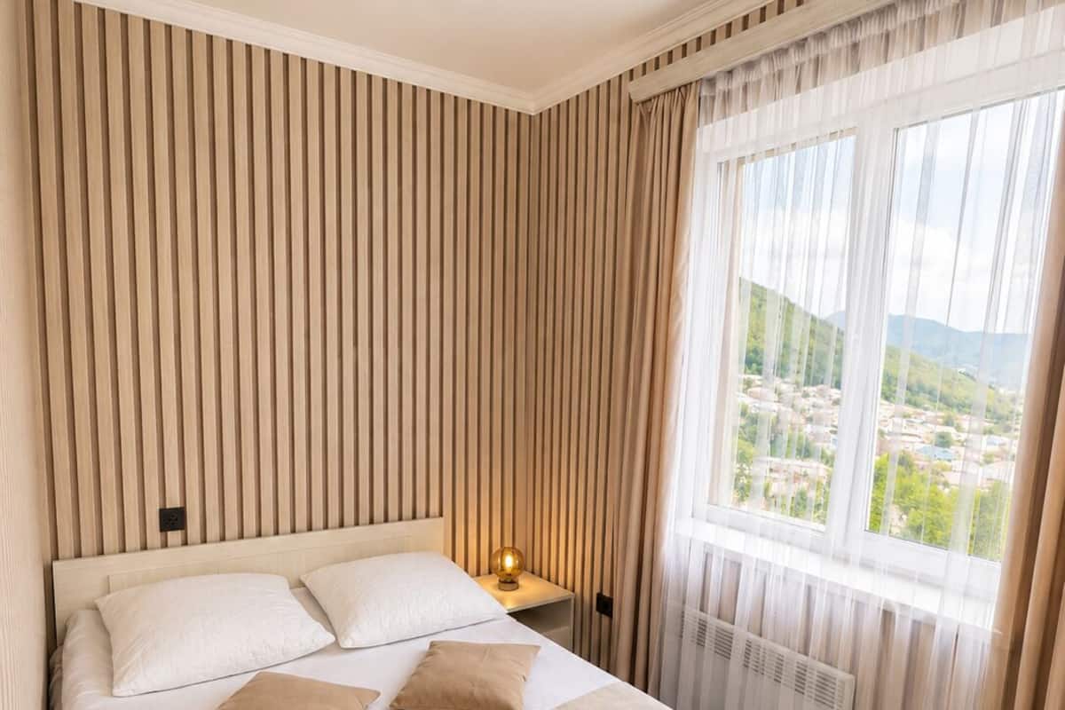 A hotel room with a large bed, a nightstand with a lamp, a window with curtains, and a view of a town.