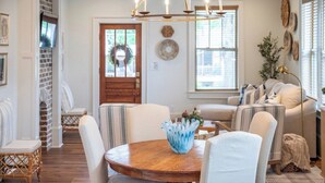 A dining area with a round wooden table, white chairs, a chandelier, and a TV mounted on the wall. - The Ole Miss Square 3BR by Velvet Ditch Villas (Oxford)