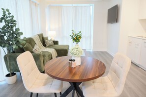 Un salón moderno con una mesa redonda de madera, dos sillas blancas, un sofá verde, una planta en maceta y un jarrón de flores.