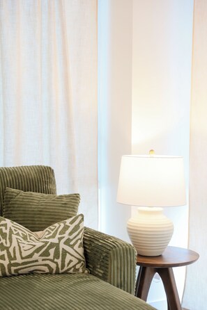 Una cama con ropa de cama verde, un cojín estampado y una lámpara blanca sobre una mesita de noche de madera.