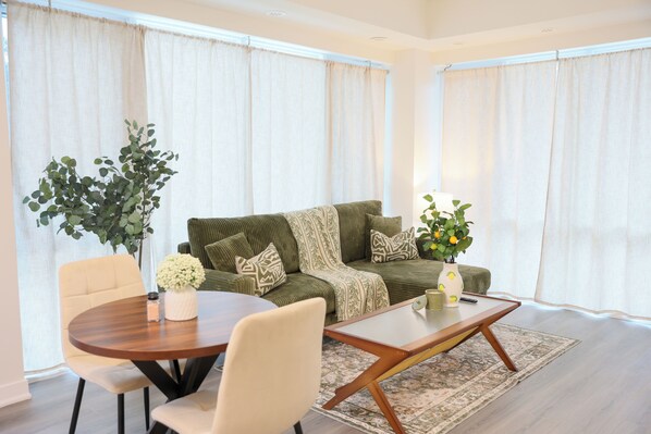 Una sala de estar con una mesa redonda de madera, sillas color beige, un sofá verde con cojines estampados y una planta en maceta.