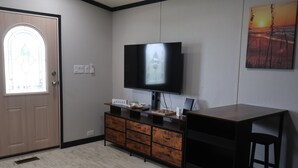 Una habitación con una televisión de pantalla plana montada en la pared, un mueble de madera con cajones, una puerta con ventanal y un cuadro en la pared.