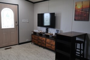 Ein Zimmer mit einem an der Wand befestigten Flachbildfernseher, einer Holzkassette mit Schubladen, einer Tür mit Glasfenster und einem Gemälde an der Wand.