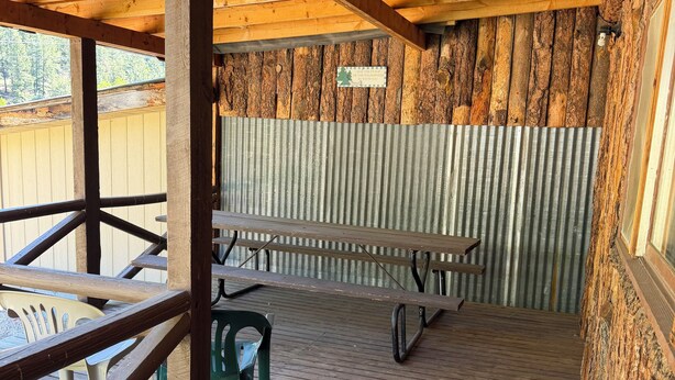 Ein überdachter Außenbereich mit Picknicktischen und Stühlen, Holzbalken und einer Wellblechwand.