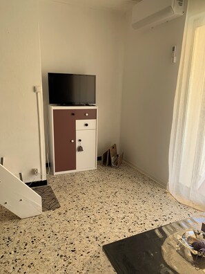 Une pièce avec un coin cuisine, un téléviseur posé sur un meuble, un tapis à motifs et une petite table avec un panier.