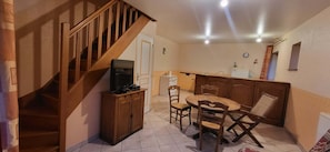 Un espace repas avec une table et des chaises en bois, un téléviseur posé sur un meuble, un escalier et, au fond, un coin cuisine.