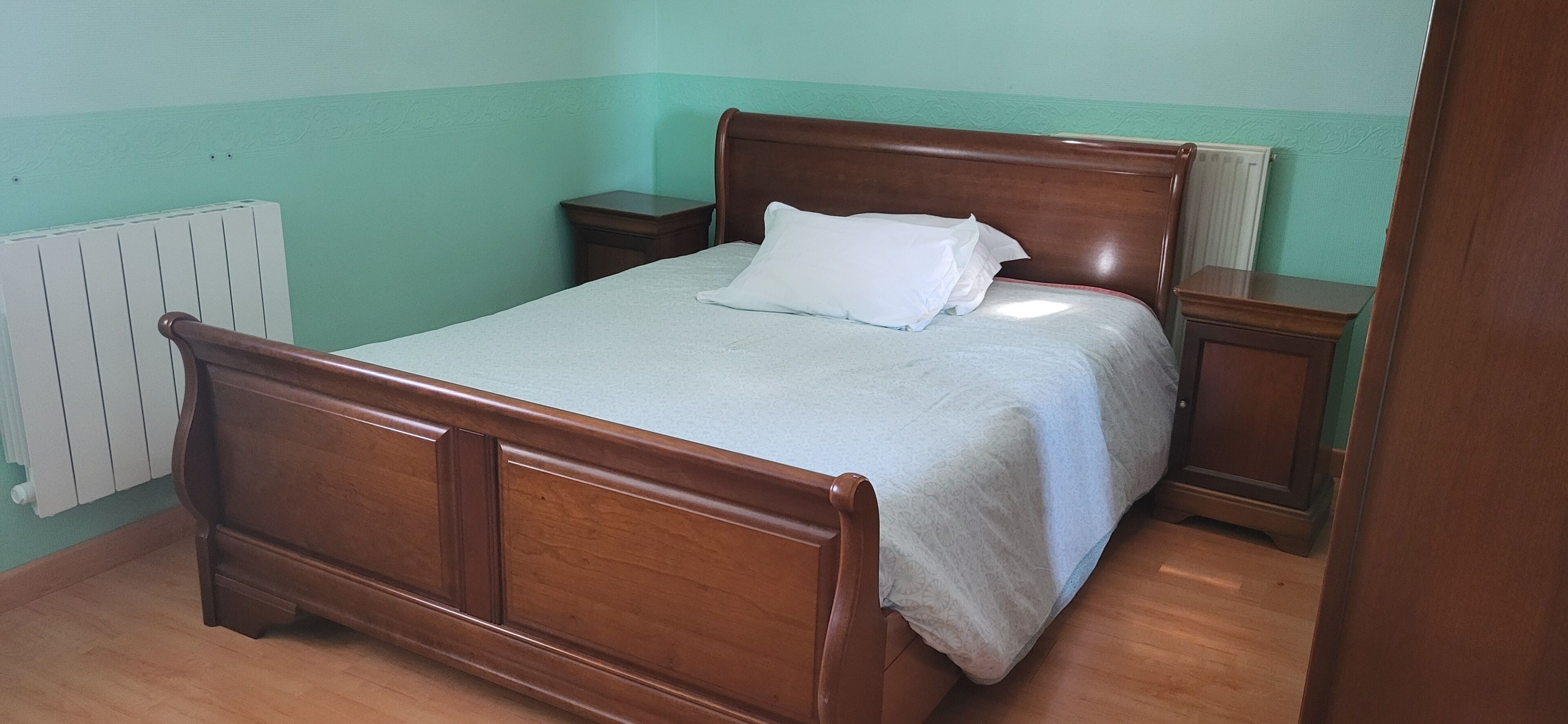 A single bed with a wooden frame, a white mattress, and a matching pillow. There is a nightstand to the right of the bed and a radiator to the left.
