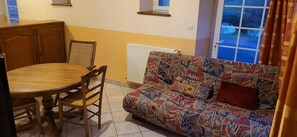 A room with a round wooden table, two chairs, a patterned sofa, a radiator, and a window with curtains.