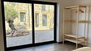 Ett rum med en stor skjutdörr i glas som leder ut till ett utomhusområde, ett trägolv, en säng med ett mönstrat överkast och en hyllsektion i trä.