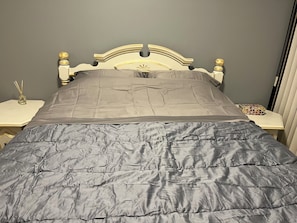 Ein Bett mit grauer Decke, einem Holzkopfschild und einem Nachttisch mit Lampe und einem Dekoartikel.
