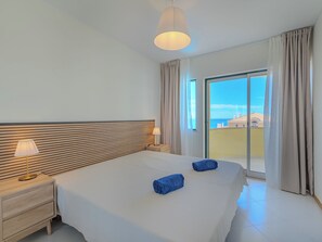 Ein Hotelzimmer mit einem großen Bett, einem Nachttisch, einer Lampe und Meerblick durch eine Glastür.