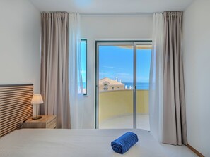 A hotel room with a bed, a nightstand, a lamp, and a view of a building and the sea through a glass door. - Algarvemor - Casa Do Tio by Interhome (Portimão)