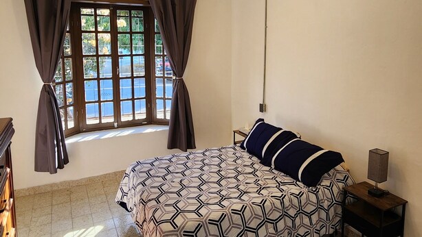 A bedroom with a bed, curtains, a nightstand, a lamp, and a ceiling light fixture. - Newly Remodeled Loft - Fast WiFi - A/C - 9 Min drive to Malecon (La Paz)