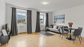 A modern living room with a sofa, dining table, and chairs, a large window with curtains, and a painting on the wall. - Faraway Homes Studios Krieglach 18 (Sommer)
