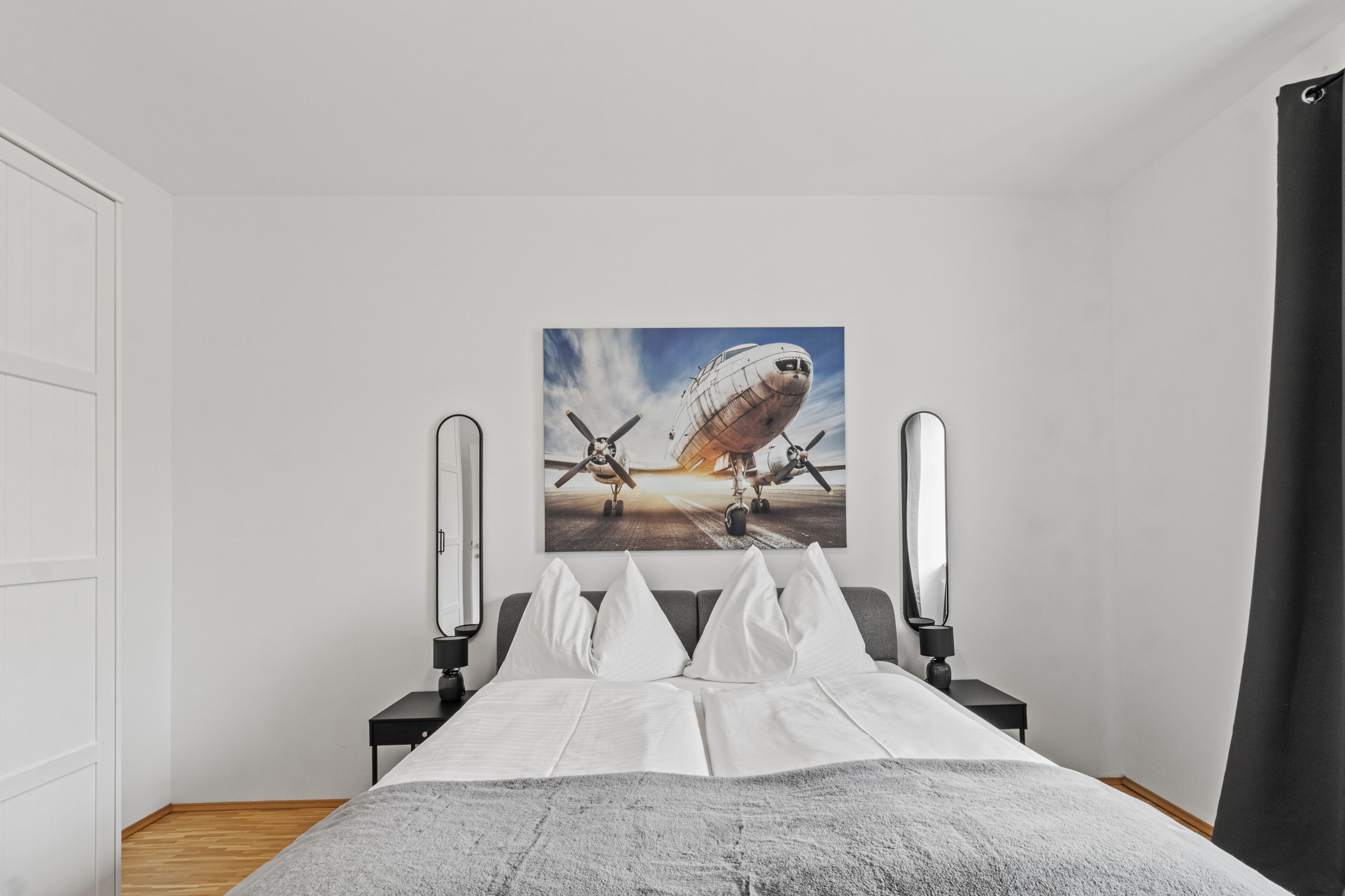 A bedroom with a bed, a nightstand, a lamp, a painting of an airplane, and a window with curtains.