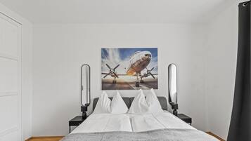 A bedroom with a bed, a nightstand, a lamp, a painting of an airplane, and a window with curtains.