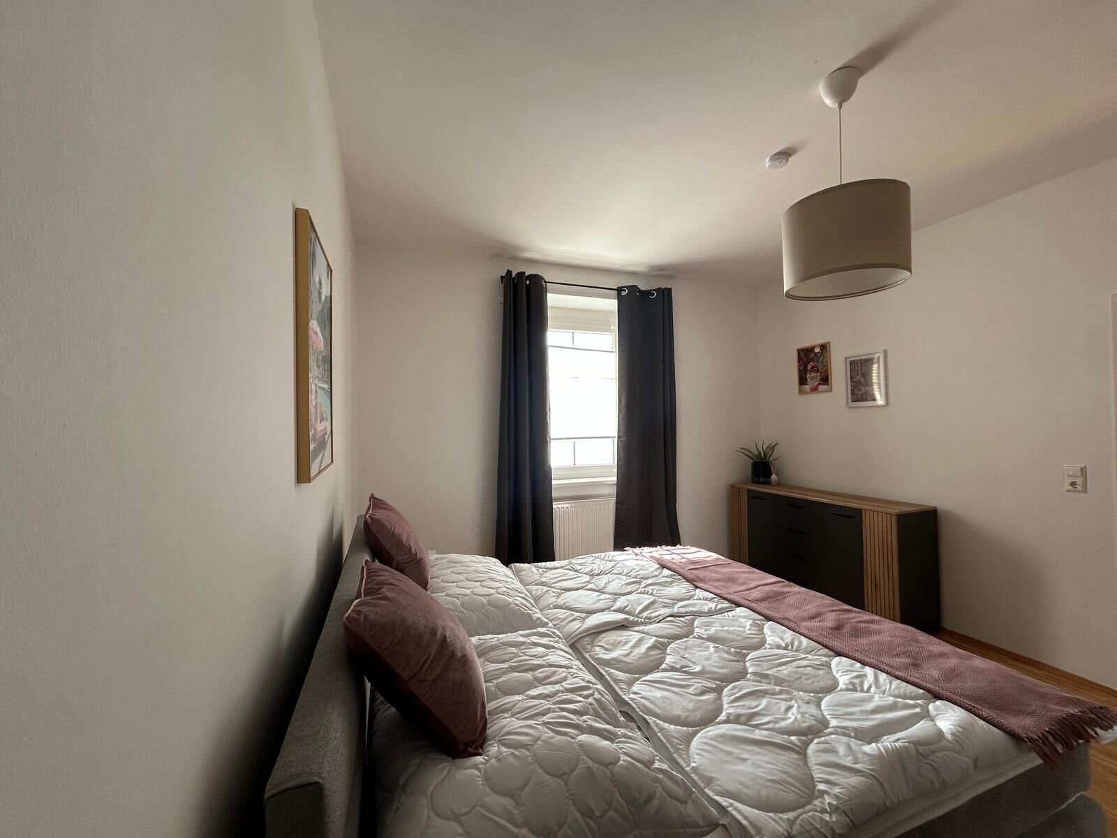 A bedroom with a bed, a window with curtains, a bedside table, and a wall-mounted lamp.