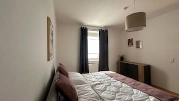 A bedroom with a bed, a window with curtains, a bedside table, and a wall-mounted lamp.
