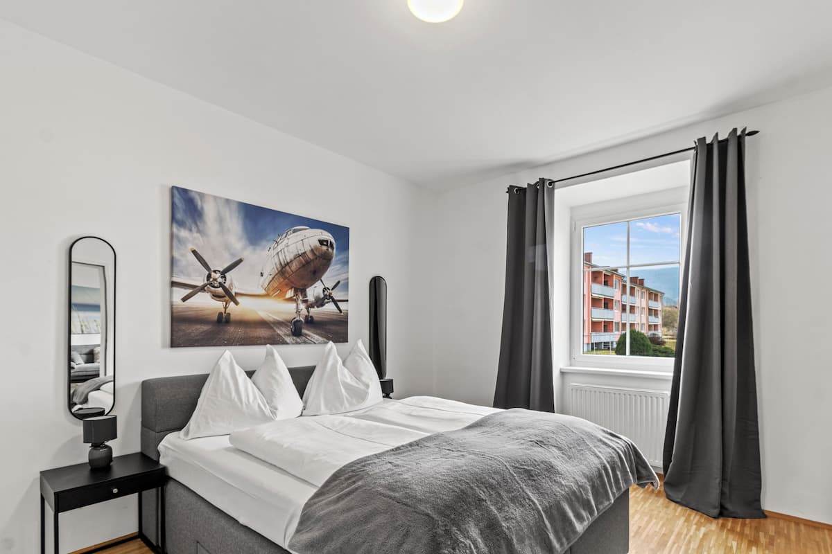 Un dormitorio con una cama, una ventana con cortinas, un cuadro de un avión y vistas de edificios desde afuera.