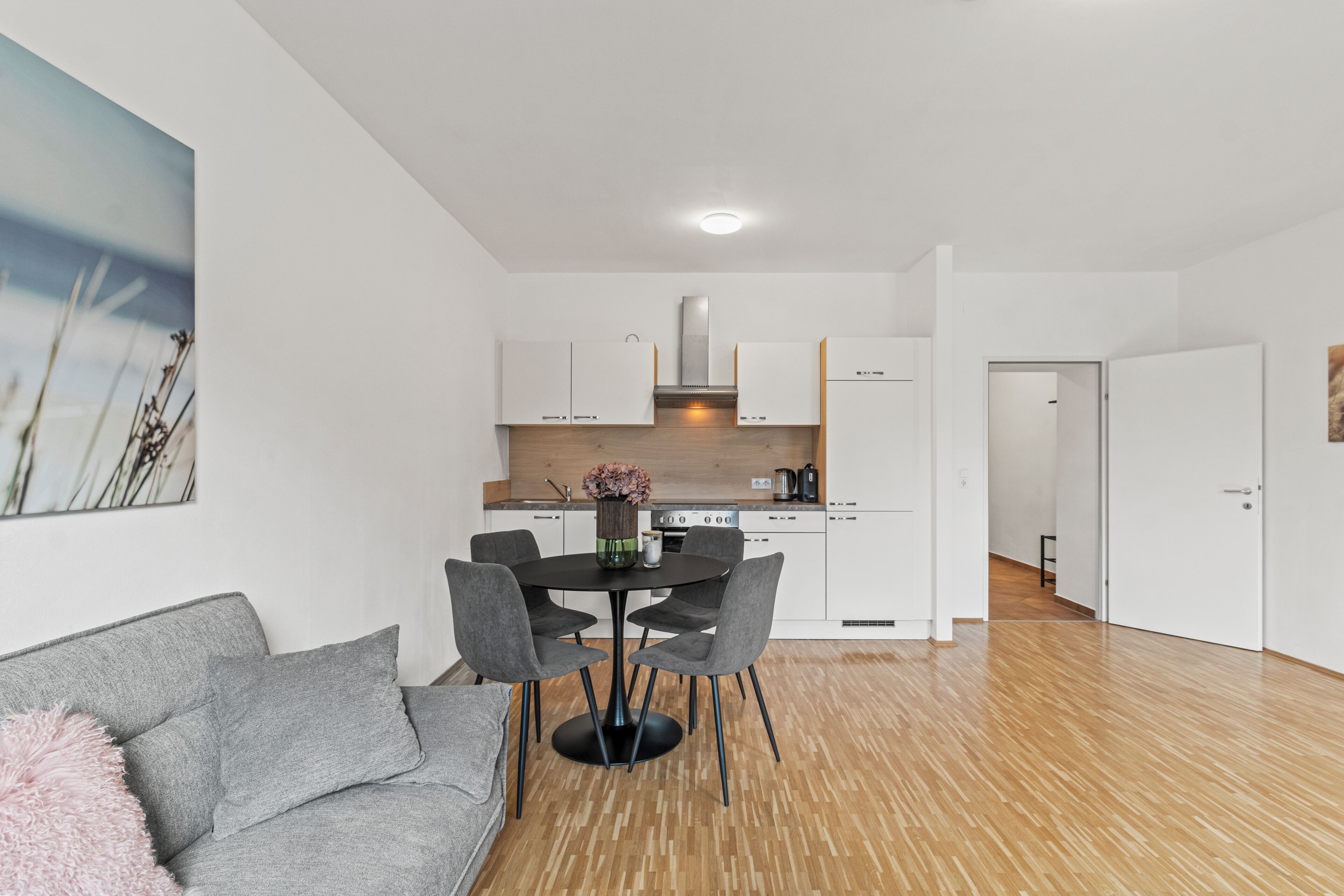 A modern kitchen with white cabinets, a central island, and a dining area with a gray sofa and a pink pillow.