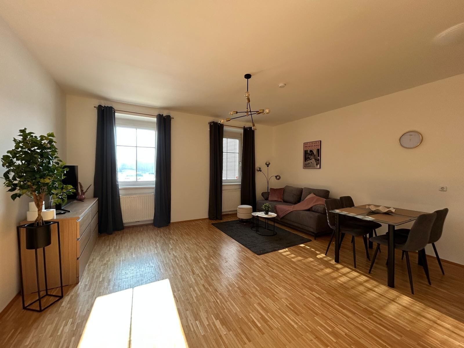 A modern living room with a sofa, dining table, and chairs, a potted plant, and a large window with curtains.