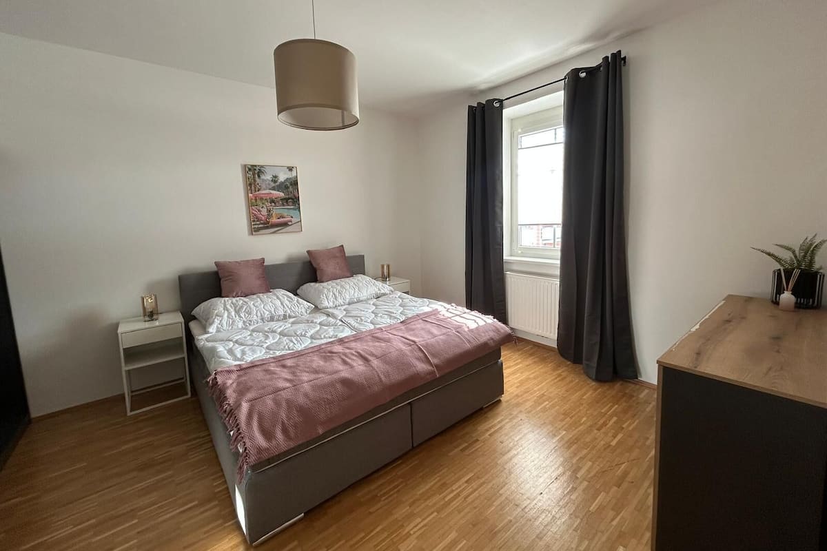 Un dormitorio con cama, mesitas de noche, lámpara colgante, ventana con cortinas y un escritorio de madera.