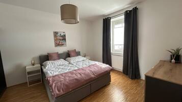 A bedroom with a bed, bedside tables, a hanging light fixture, a window with curtains, and a wooden desk.