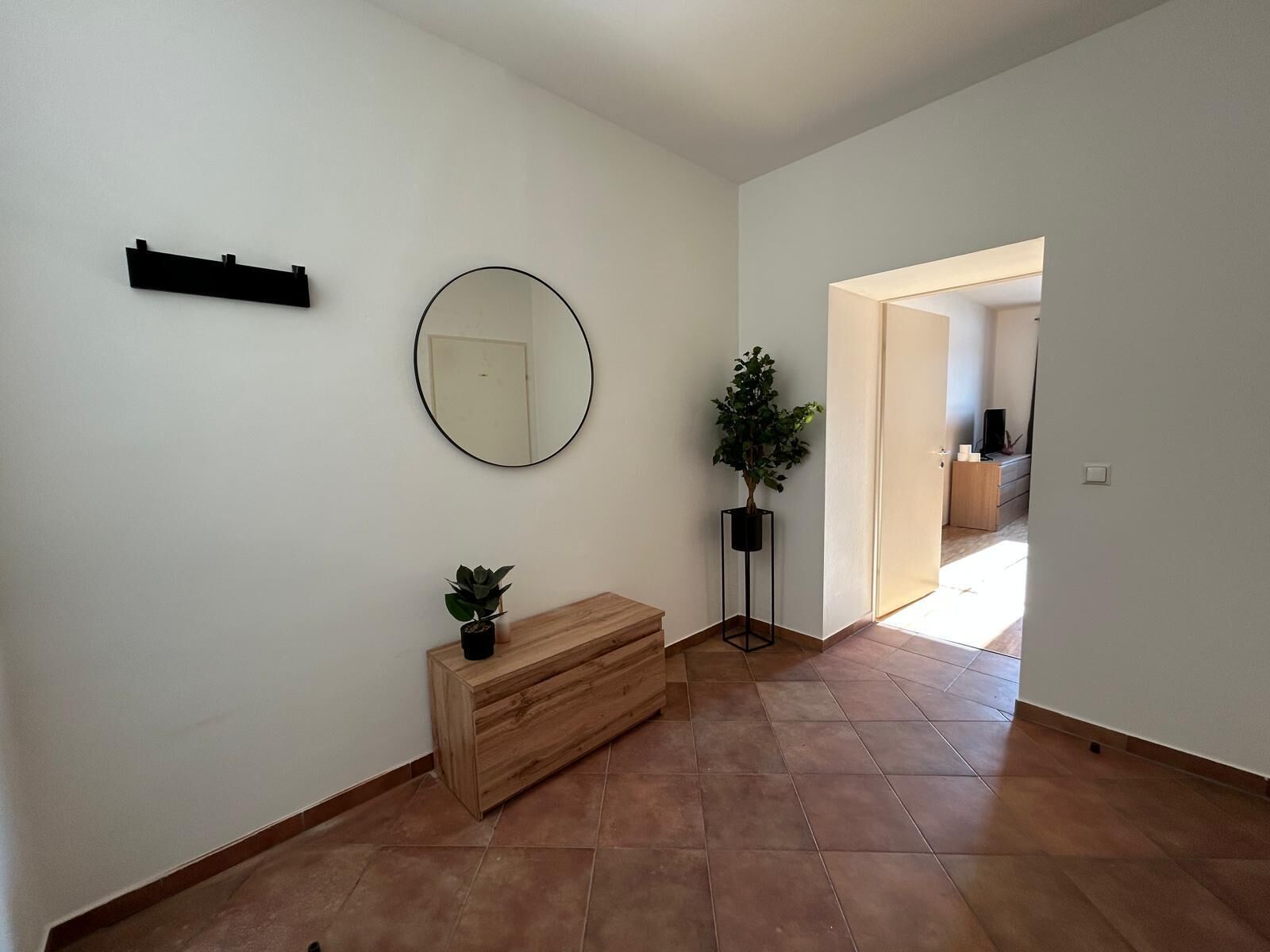 A hallway with a wooden bench, a round mirror, a potted plant, and a door leading to another room.