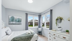 A bedroom with a bed, a chair, a dresser, a window with curtains, and a painting on the wall. - Cozy Cityscapes 3Br, 1B with Washer+Dryer (St. Catharines)