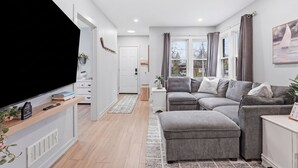 A modern living room with a flat-screen TV, grey sofas, a white side table, and a large window with curtains. - Cozy Cityscapes 3Br, 1B with Washer+Dryer (St. Catharines)
