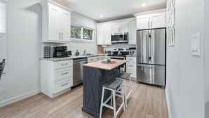 A modern kitchen with white cabinets, stainless steel appliances, a central island with stools, and a window. - Cozy Cityscapes 3Br, 1B with Washer+Dryer (St. Catharines)
