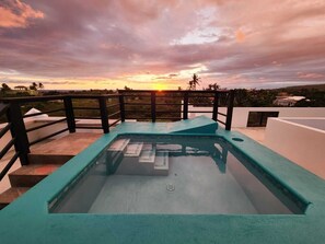 A swimming pool with a glass bottom, surrounded by a wooden deck and a metal railing, with a sunset view in the background.
