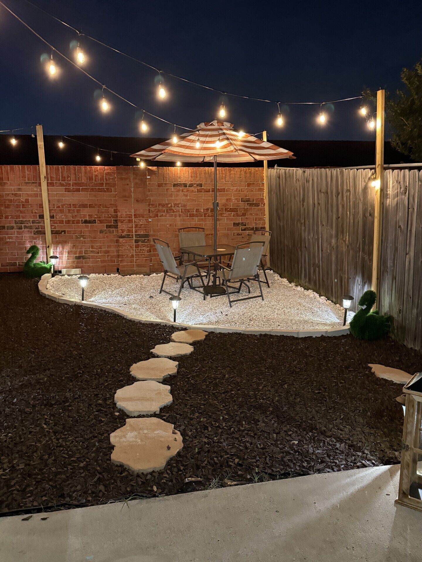 A patio with a brick wall, a wooden fence, a string of lights, a table with chairs, and a decorative swan statue.
