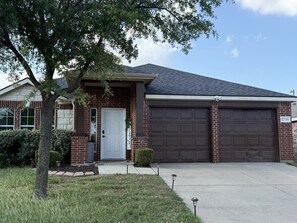 A residential house with a brick facade, a front door, two garage doors, and a well-maintained lawn. - Modern Comfort in the Heart of Texas (Grand Prairie)