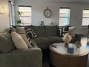 Ein Wohnzimmer mit einer Eckcouch, einem runden Couchtisch, einer Wanduhr und einem Fenster mit Jalousien.