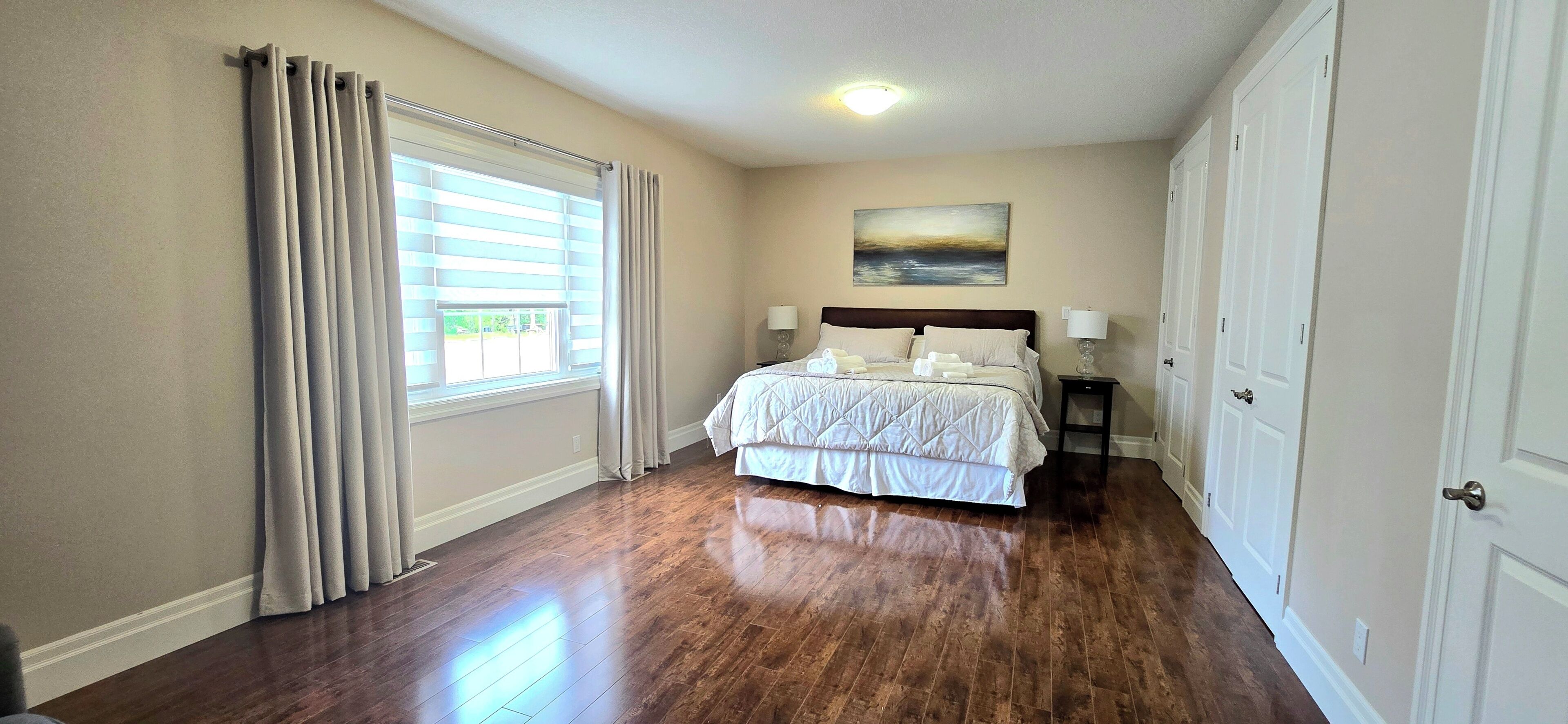 A bedroom with a large bed, a window with blinds, a painting on the wall, and a door on the right.