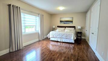 A bedroom with a large bed, a window with blinds, a painting on the wall, and a door on the right.