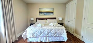 A neatly made bed with a quilted duvet, two bedside lamps, a painting on the wall, and a door to the right.