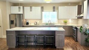 A modern kitchen with a central island, bar stools, stainless steel appliances, and a window providing natural light.